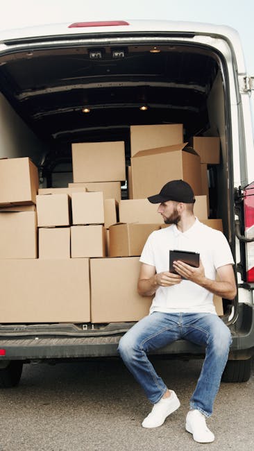 A young male mover with long hair, wearing a black bandana and dark clothing, is sitting in the driver's seat of a white moving van decorated with the company's name 'MOVING COMPANY' and the words 'LO