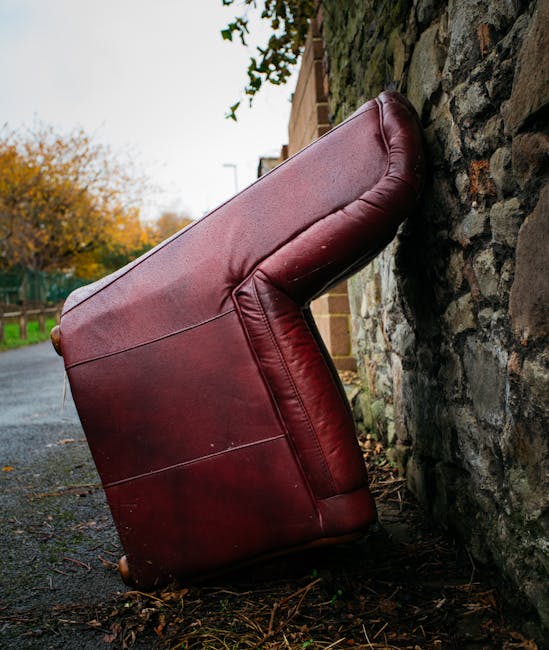 A large, burgundy leather armchair is shown leaning against a rough stone wall on a paved outdoor driveway, partially outside a residential property. The chair is positioned on its side, with the backrest and armrest visible, and appears to be in the process of being moved or discarded as part of a home relocation or furniture disposal. Surrounding the chair are scattered leaves and small debris, with a background featuring a brick building, a wooden fence, and trees with autumn foliage under an overcast sky. The scene is illuminated by natural daylight, suggesting daytime conditions suitable for loading and transport activities typically associated with house removals. The image reflects the type of furniture transport and packing processes that Pimlico Man and Van may handle when disposing of bulky items in line with local council rules for furniture disposal in Pimlico.