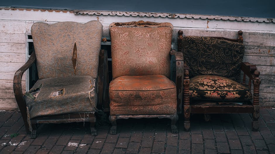 Three old, worn-out armchairs placed side by side outdoors on a brick pavement in front of a weathered white wooden wall. The first armchair on the left has torn fabric upholstery, exposing the cushioning and frame beneath, with visible holes and frayed edges. The middle armchair features faded, patterned fabric with signs of age and slight discoloration, and its wooden legs are intact. The third armchair on the right has dark, ornate upholstery with intricate gold and red patterns, supported by turned wooden arms and legs, and appears to be in slightly better condition. All three chairs are positioned close together, likely awaiting removal or disposal as part of house clearance or furniture transportation services. The setting suggests an outdoor space near a property, possibly during a home relocation or furniture disposal process, with natural lighting highlighting the textures and wear of each piece, illustrating a typical scene for furniture removal tasks handled by Pimlico Man and Van.
