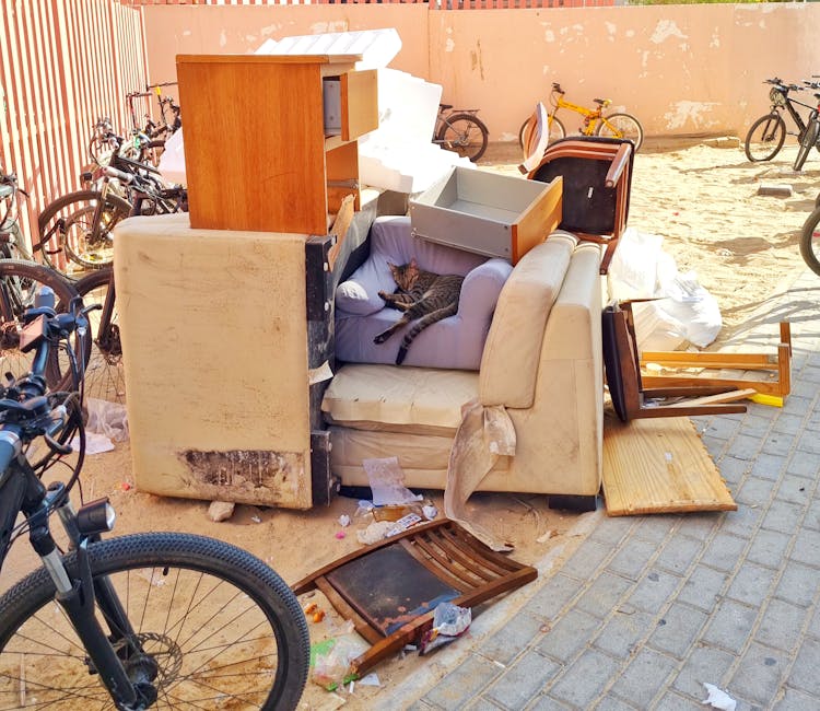 A large, burgundy leather armchair is shown leaning against a rough stone wall on a paved outdoor driveway, partially outside a residential property. The chair is positioned on its side, with the backrest and armrest visible, and appears to be in the process of being moved or discarded as part of a home relocation or furniture disposal. Surrounding the chair are scattered leaves and small debris, with a background featuring a brick building, a wooden fence, and trees with autumn foliage under an overcast sky. The scene is illuminated by natural daylight, suggesting daytime conditions suitable for loading and transport activities typically associated with house removals. The image reflects the type of furniture transport and packing processes that Pimlico Man and Van may handle when disposing of bulky items in line with local council rules for furniture disposal in Pimlico.