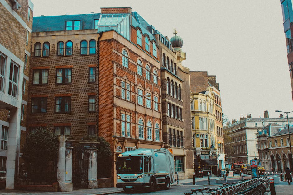 A white pallet truck equipped with a flat platform and safety straps is positioned on a paved street in front of a multi-storey building with ornate windows and a brick facade. The vehicle is loaded with large cardboard boxes, some wrapped in plastic, supporting a home relocation or furniture transport process. The scene is set in an urban area with a mixture of historic and modern architecture, and several other vehicles, including a small van, are parked nearby. Adjacent buildings feature detailed architecture with arched windows, decorative stonework, and a variety of exterior materials such as brick and stucco. The street is lined with black bollards, and adjacent to the sidewalk are bike racks and a lamppost, indicating a busy city environment suitable for professional removals like those offered by Pimlico Man and Van. The lighting suggests a clear day, ideal for loading and moving activities related to property relocation on Grosvenor Road, Pimlico.