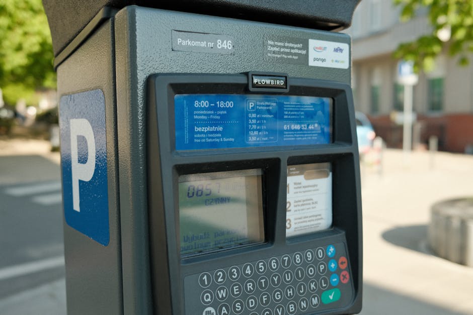 Close-up image of a parking meter situated outdoors on a city street, with a blue 'P' sign indicating parking. The parking meter displays a screen showing the current parking fee rates and hours of operation, with details such as '8:00 – 18:00, Monday – Friday' and free parking on Saturdays and Sundays. The meter is made of dark grey metal and features a digital display, keypad, and card slot for payment. In the background, blurred elements of a street scene are visible, including a tree with green leaves, nearby residential buildings, and parked cars. The parking meter is part of a home relocation or furniture transport process managed by Pimlico Man and Van, supporting efficient moving logistics and parking permit management for house removals in Pimlico.
