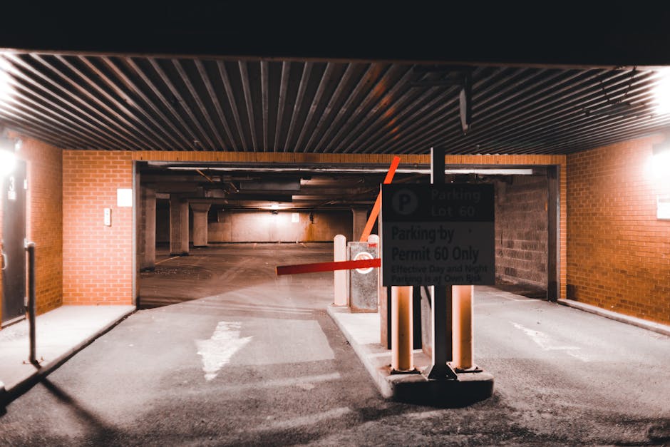 An underground parking garage entrance with a barrier gate, featuring a red arm and a black and white pole, located within a brick building structure. A sign attached to the barrier displays parking regulations specific to parking lot 60 and permit requirements. Inside the garage, the concrete floor extends into the dimly lit internal space, with visible support columns and a garage ceiling with wooden or metal slats. The area is illuminated by artificial lighting on the right side, and a painted arrow on the pavement indicates the direction of vehicle movement. Adjacent to the entrance, there are brick walls and some equipment that might be related to parking management or security. The scene captures a typical setting for vehicle access control in a residential or commercial property involving home relocation or furniture transport logistics, as managed by a professional removals company like Pimlico Man and Van.