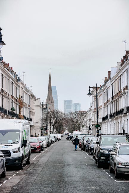 A wide view of a residential street in Pimlico, London, lined with white terraced houses featuring black wrought iron balconies and window railings. Parked cars, including a white van used by Pimlico Man and Van, line both sides of the street. In the foreground, a person dressed in dark clothing is carrying a cardboard box, preparing for home relocation or furniture transport. Behind, a moving trolley and packing materials such as plastic wrapping and blankets are visible, indicating a loading process. The street leads towards a historic church with a tall, pointed spire and modern high-rise buildings in the background, under an overcast sky. The scene captures a typical urban environment suitable for removals and moving services in SW1V, with the presence of professional transport equipment and clear activity relevant to packing and moving logistics.