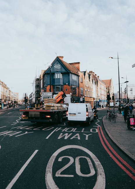 A white flatbed delivery truck with an open bed and black chassis is parked on a residential street in Pimlico, SW1V, during daytime. The truck is loaded with wooden pallets wrapped in plastic and other packing materials, ready for home relocation. Nearby, a white panel van is parked alongside the truck, with its rear doors closed. The street features a marked speed limit of 20 miles per hour, with red double lines along the curb indicating parking restrictions. In the background, there are multi-storey brick and timber residential buildings with pitched roofs, and the sky above is partly cloudy. On the pavement, a bicycle is parked upright, and pedestrians are visible walking along the sidewalk. Streetlights and traffic signals are positioned along the road, supporting the logistics and transportation process involved in house removals. The scene depicts the loading and transportation phase typical of furniture transport and packing and moving services offered by Pimlico Man and Van.