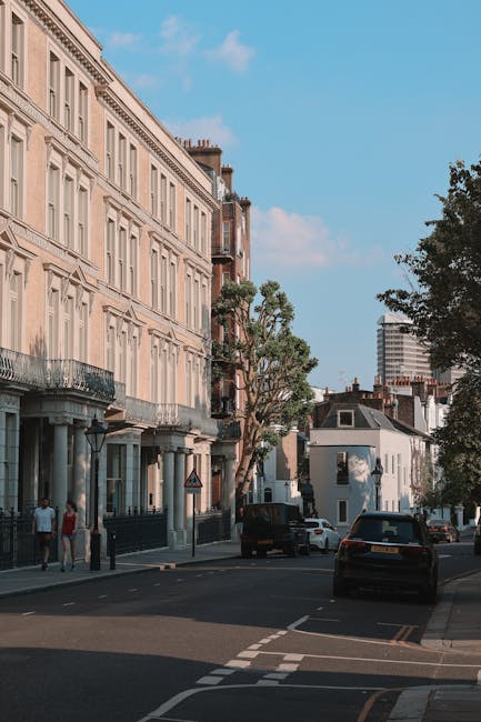 A wide view of a residential street in Pimlico, London, lined with white terraced houses featuring black wrought iron balconies and window railings. Parked cars, including a white van used by Pimlico Man and Van, line both sides of the street. In the foreground, a person dressed in dark clothing is carrying a cardboard box, preparing for home relocation or furniture transport. Behind, a moving trolley and packing materials such as plastic wrapping and blankets are visible, indicating a loading process. The street leads towards a historic church with a tall, pointed spire and modern high-rise buildings in the background, under an overcast sky. The scene captures a typical urban environment suitable for removals and moving services in SW1V, with the presence of professional transport equipment and clear activity relevant to packing and moving logistics.