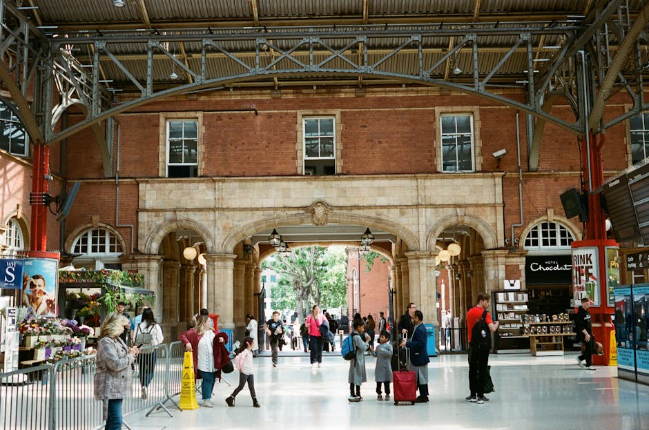 Inside a busy train station with a high, arched ceiling made of metal and glass, supported by red-painted steel beams. The station features a large, ornate stone archway with decorative carvings, leading to an outdoor area where trees and natural light are visible. The platform area includes a mix of people, some carrying luggage or backpacks, and others waiting or walking. On the left side, there is a flower stall with a variety of plants and flowers displayed on shelves, and a woman wearing glasses and a grey coat is seen looking at the flowers. Near the center, a family with children and a person with a luggage trolley are engaged in conversation or moving through the station. To the right, there is a small kiosk selling snacks and souvenirs, with personnel attending to customers. The scene captures the typical atmosphere of a London station involved in home relocation or transit, with visible movement, seating, and signage, suitable for a house removals or furniture transport context supported by Pimlico Man and Van.