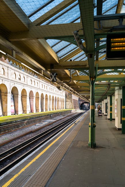 A view of Pimlico Station platform showing multiple sets of railway tracks running parallel along the length of the station. The platform is partially covered by a metal and glass roof supported by green-painted metal columns and frameworks. Along the station wall, there are brick arches and advertising posters, with some posters visible on the left side of the image. The platform surface is made of concrete, featuring tactile paving strips near the edge for safety. In the background, the station tunnel is visible, and a few passengers with luggage and shopping bags can be seen waiting or walking along the platform. This station is part of an urban rail network, with clear signage and lighting highlighting the area. The setting is well-lit by natural daylight filtering through the roof panels, and the overall scene portrays a typical train station environment suitable for home relocation or furniture transport within urban moving services offered by Pimlico Man and Van.