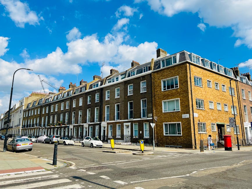 A street view of a multi-storey residential building constructed with brown bricks, featuring large bay windows and rooftop dormer windows under a partly cloudy blue sky. In the foreground, a parking area with several parked cars is visible, along with a sidewalk where a person is walking past a red post box. A lamppost stands on the pavement, and the area appears to be part of an urban neighbourhood. This scene provides context for house removals and furniture transport services, illustrating the typical exterior environment involved in home relocation projects handled by Pimlico Man and Van. The setting is well-lit during daytime, with no visible moving activity in progress but suggests a possible location for loading or unloading for a nearby move.