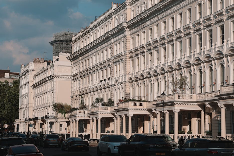 A row of white, multi-storey Victorian-style terraced houses with ornate architectural details such as arched windows, decorative cornices, and balconies with balustrades, situated along a street in Pimlico. The buildings are closely spaced, with some featuring potted plants on balconies or window sills, and vehicle parking along the curb. The street scene shows parked cars and street lamps, with a partly cloudy sky overhead. This setting reflects a typical residential area where professional house removals and furniture transport, such as those provided by Pimlico Man and Van, may be required for home relocation or moving services, involving the careful handling and loading of household items into vans for transportation.