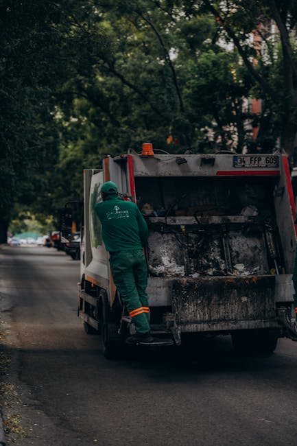 A sanitation worker wearing a green uniform and protective helmet is seen standing on the back step of a rubbish collection vehicle on a residential street lined with trees. The vehicle's rear compartment is open, revealing accumulated debris and waste bags. The worker appears to be in the process of loading or securing waste materials, with the vehicle positioned on the tarmac road adjacent to parked cars and a sidewalk. In the background, there are leafy branches overhead and multi-storey buildings partially visible through the trees. The scene takes place during daylight, with natural light illuminating the surroundings. This image, captured as part of home relocation or waste collection services, highlights the logistical process involved in clearing bulky household waste, an essential step in move-related clean-up activities managed by Pimlico Man and Van.