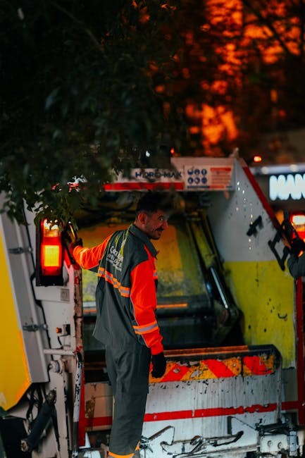 A worker wearing a high-visibility jacket with orange and reflective stripes is standing next to a large waste collection vehicle on a street at dusk. The vehicle, which appears to be used for bulky waste or debris collection, has its rear loading area open, revealing empty compartments with protective plastic and metal surfaces. The worker is holding onto the side of the vehicle, possibly preparing to load or unload items, with his other hand resting on the vehicle's edge. Behind him, the vehicle’s lights are illuminated, and the background shows a dark sky with orange hues from sunset and faint silhouettes of trees. The scene suggests a street-level waste removal operation consistent with house clearance or moving logistics. This image conveys the professional handling of waste as part of a home relocation process, highlighting efficient logistics that a removals specialist like Pimlico Man and Van might undertake during furniture transport or packing and moving services.